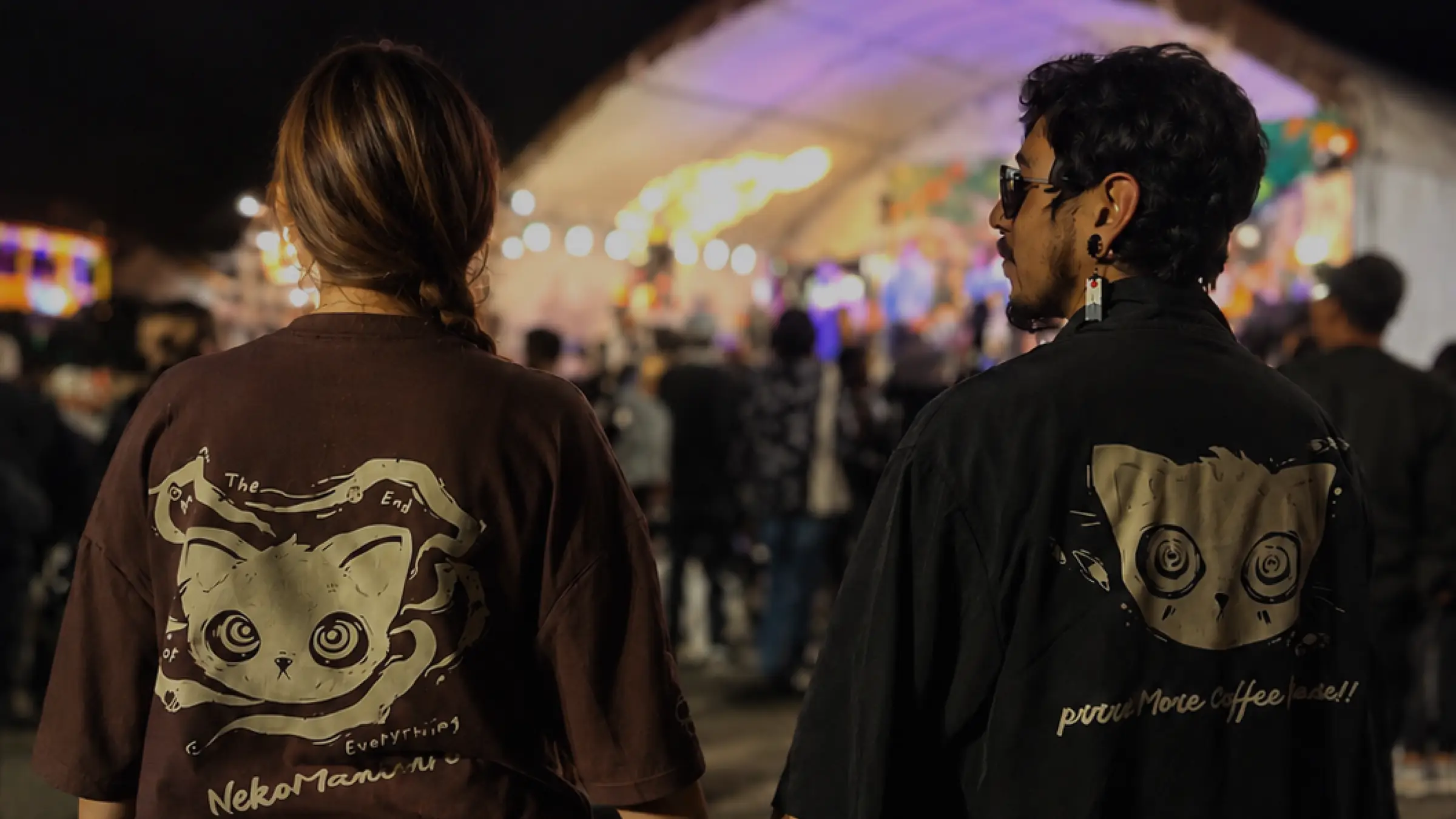 Dos personas de espaldas en un festival nocturno usando camisetas Malsano con el gato de ojos en espiral